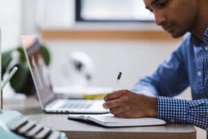a person writing on a notebook sitting in front of a laptop
